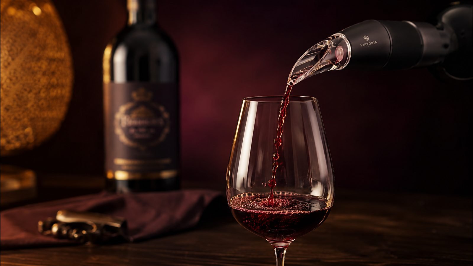 A glass of red wine being poured from an automatic wine dispenser, with a corkscrew and a bottle of red wine in the background on a wooden table. The scene is dimly lit with a warm, elegant atmosphere.