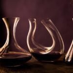Three glass wine decanters on a dark timber table: a wide-based crystal decanter with red wine, a curved sculptural decanter and a simple tall decanter
