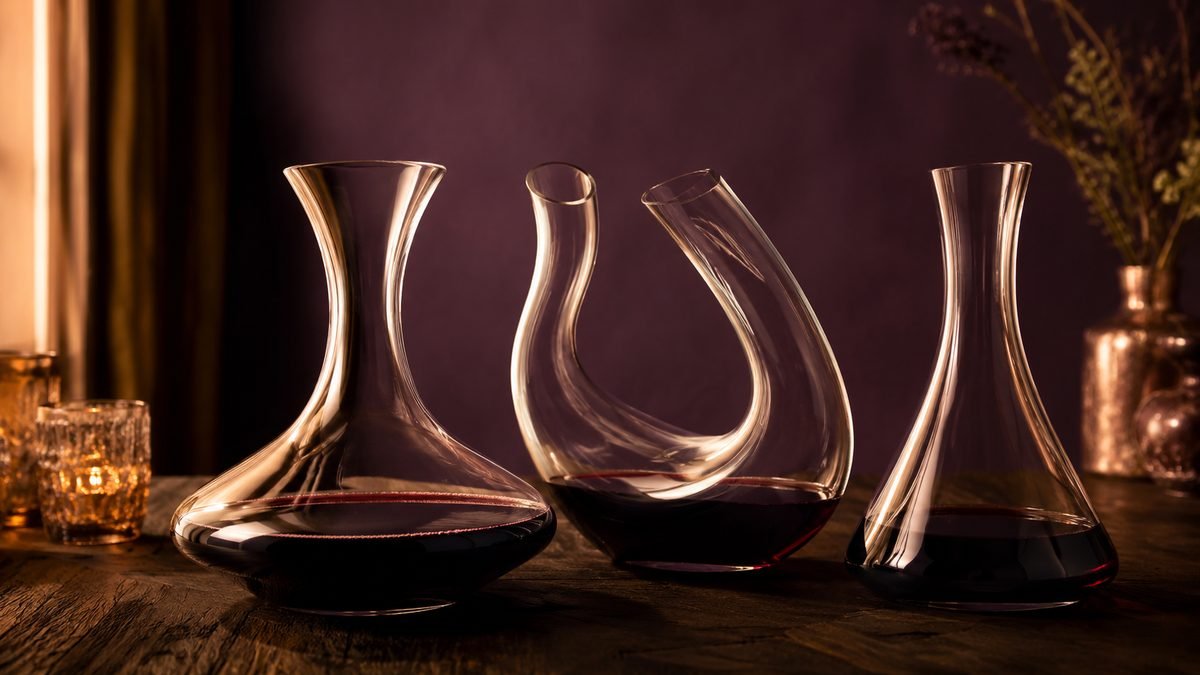 Three glass wine decanters on a dark timber table: a wide-based crystal decanter with red wine, a curved sculptural decanter and a simple tall decanter