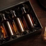 An opened premium gift box on a dark walnut table containing three small amber spirit bottles in dark tissue paper, with a glass of whisky beside it in warm moody lighting.