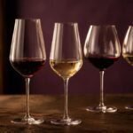 Four elegant wine glasses on a dark timber table: two stemmed crystal glasses with red and white wine and a matching set of universal glasses behind them